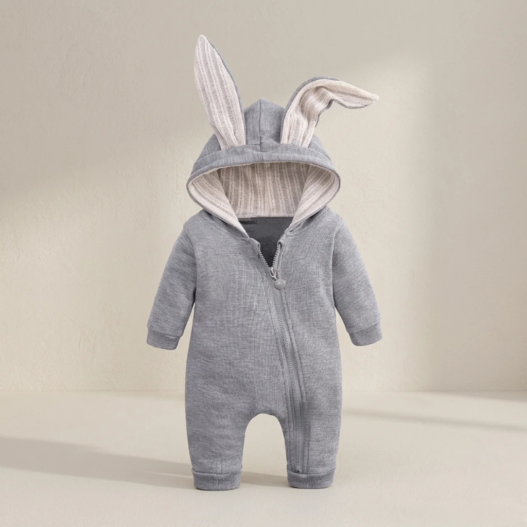 A baby hooded one-piece romper in heather gray fabric with plush bunny ears on the hood, featuring a front zipper and ribbed trim inside the hood, shown against a warm beige studio background.