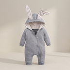 A baby hooded one-piece romper in heather gray fabric with plush bunny ears on the hood, featuring a front zipper and ribbed trim inside the hood, shown against a warm beige studio background.