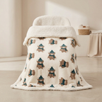 A plush cream-colored blanket draped over a chair in a softly lit room. The blanket features an allover pattern of teddy bears wearing teal hats and brown scarves, with a thick, soft fleece-like cream border.
