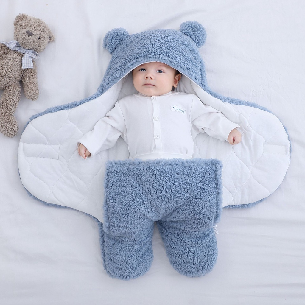 Baby in a blue bear-themed outfit lying on a white blanket with a teddy bear beside.