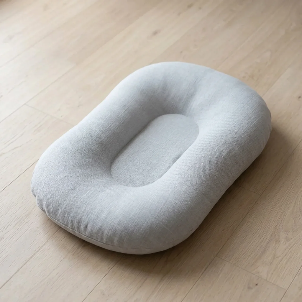 A light gray fabric baby support pillow with an ergonomic contour design, resting on a light wood floor.