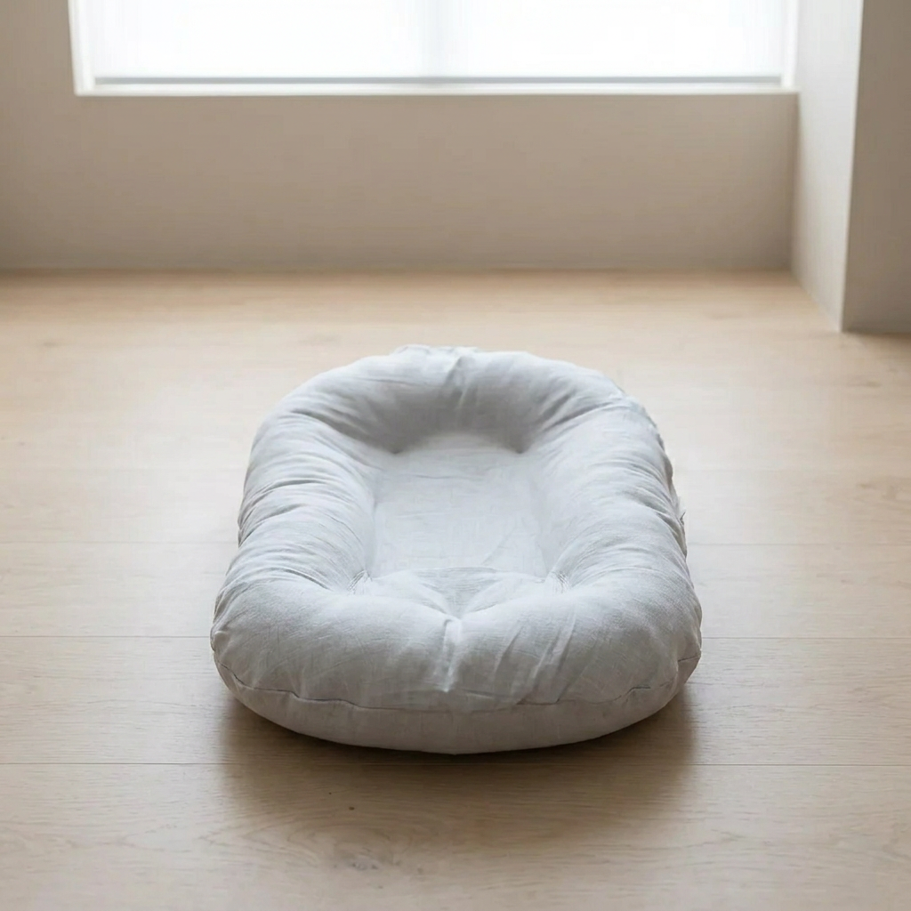 A light gray fabric baby lounger or cushion with a tufted center, placed on a light wood floor in a minimalist room with a window in the background.
