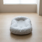 A light gray fabric baby lounger or cushion with a tufted center, placed on a light wood floor in a minimalist room with a window in the background.