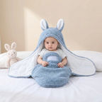 A baby sits wrapped in a plush blue bunny hooded blanket with white inner lining, photographed on a white bed with a stuffed bunny toy beside them.