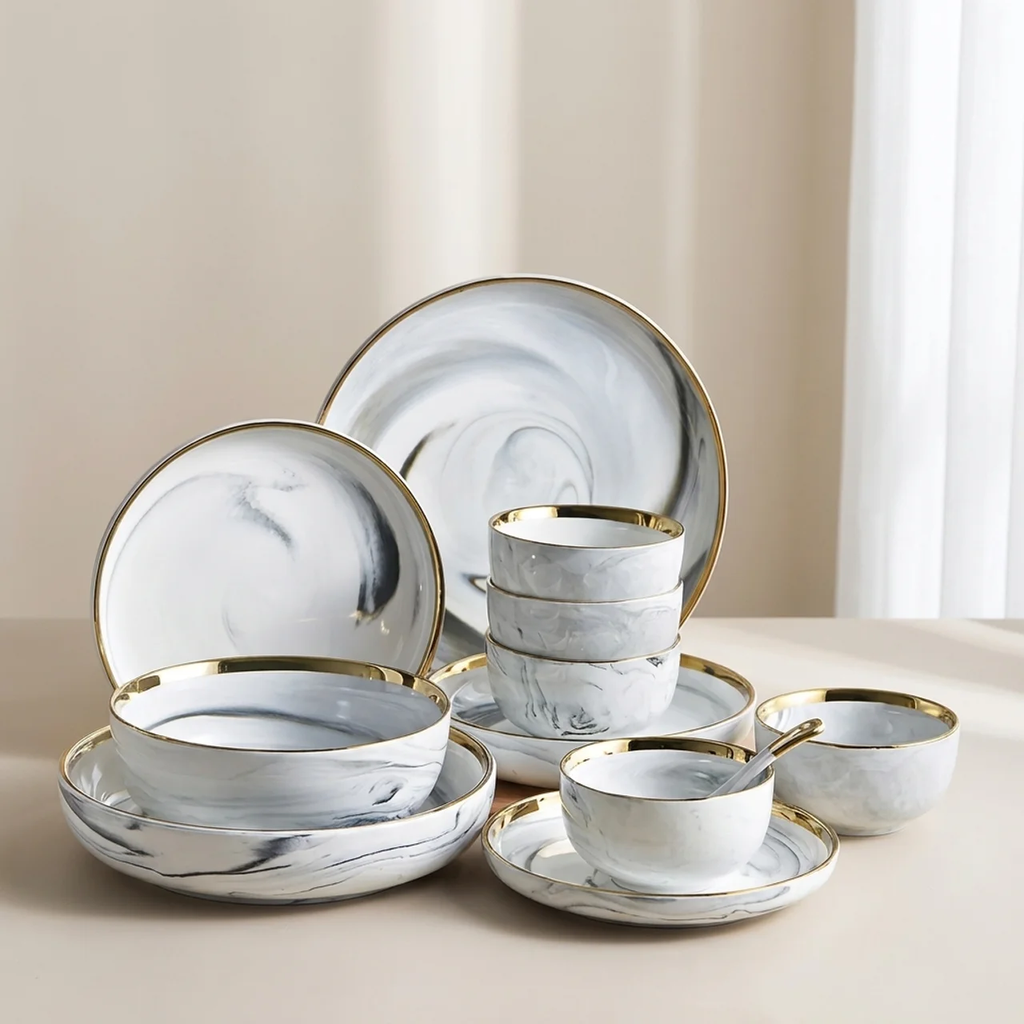 A coordinated dinnerware set with white and gray marbled plates, bowls, plates, and cups, each accented with gold trim, arranged on a light beige tabletop against a soft neutral background.