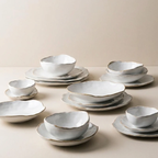 A coordinated set of ceramic dinnerware in a soft off-white glaze with subtle gold-toned edging, arranged on plates against a warm beige studio background.