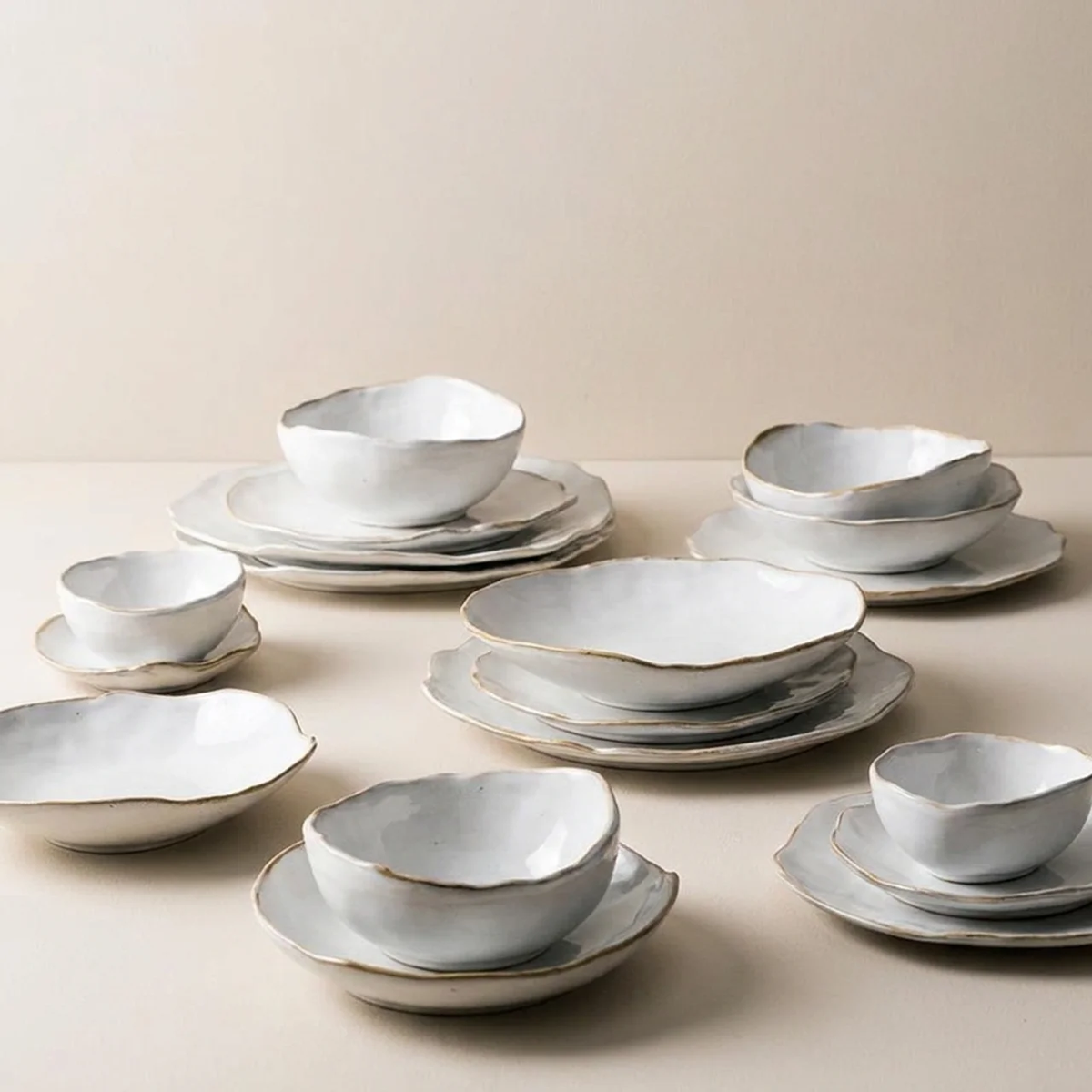 A coordinated set of ceramic dinnerware in a soft off-white glaze with subtle gold-toned edging, arranged on plates against a warm beige studio background.
