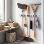 A wall-mounted broom and mop storage rack in a bright bathroom or utility room, holding several cleaning tools including a broom, squeegee, dustpan, scrubber, and towel. A wooden shelf, basket, and laundry hamper are visible nearby.