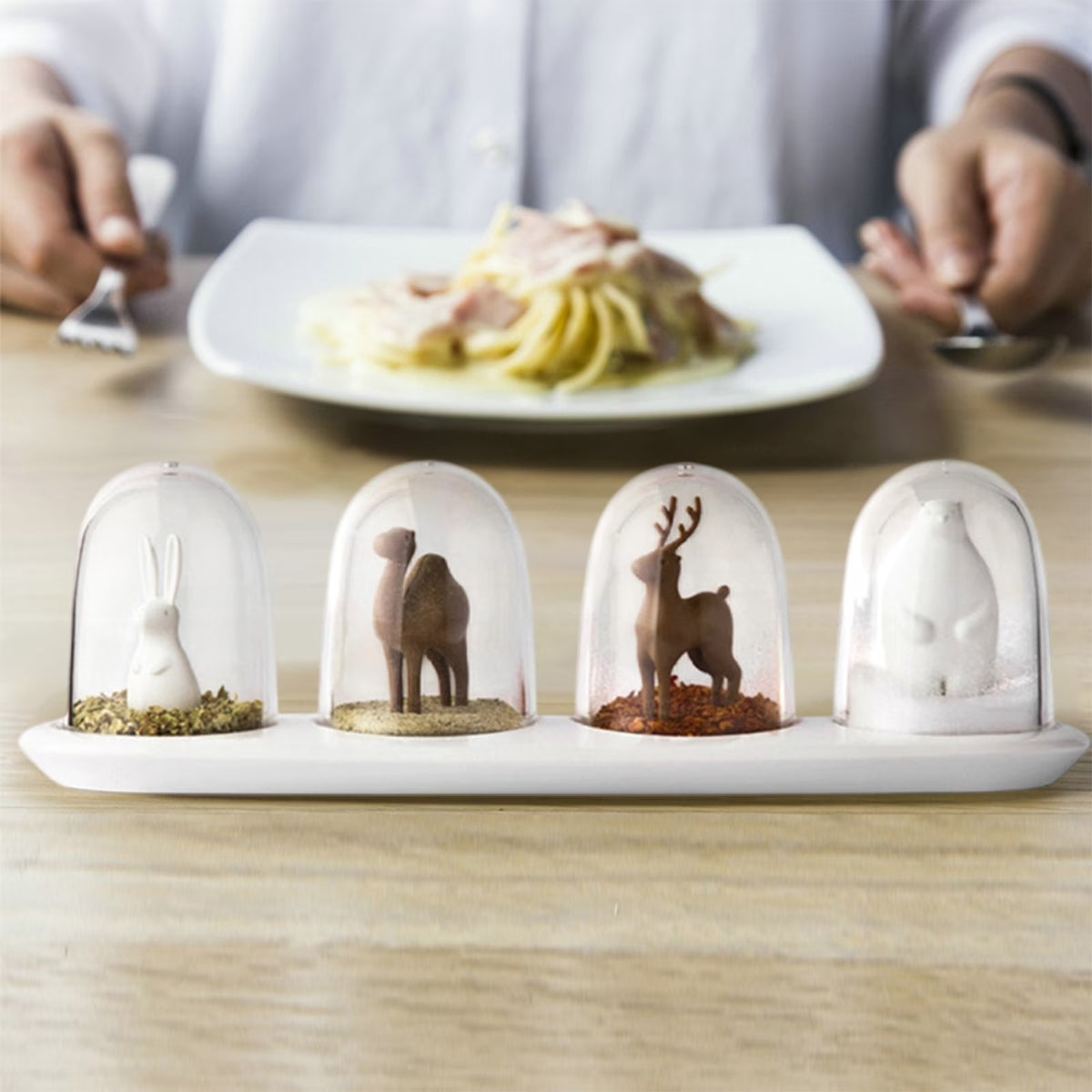 Four glass dome ornaments with miniature animal figurines on a white tray, with pasta, plate, fork, and spoon in the background.