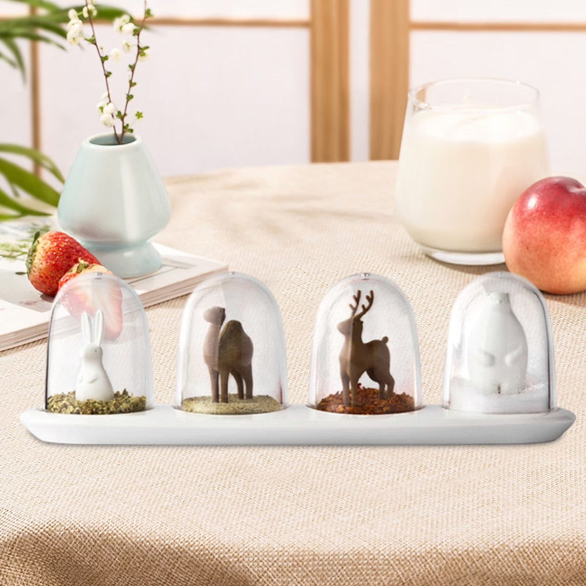 Four glass dome ornaments with miniature animal figurines on a white tray, with a vase, milk, strawberries, and peach in the background.