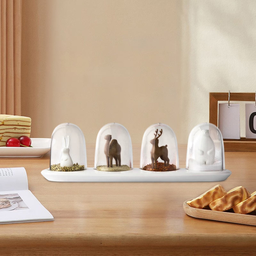 Four glass dome ornaments with miniature animal figurines on a white tray, with cake, cherries, pastries, framed decor, and an open magazine in the background.