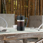 French press with coffee on a table outdoors, surrounded by bamboo and a chair.