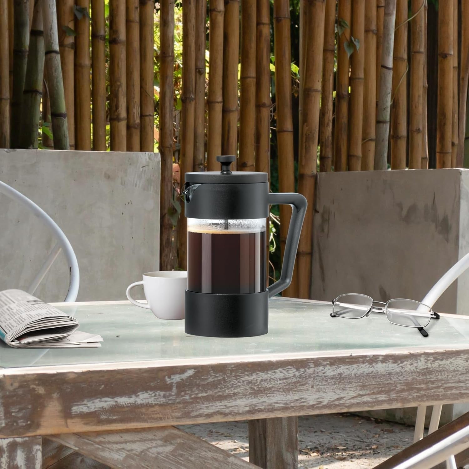 French press with coffee on a table outdoors, surrounded by bamboo and a chair.