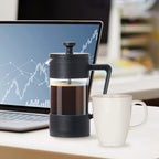 Black French press filled with coffee next to a white mug on a desk with a laptop displaying financial charts.