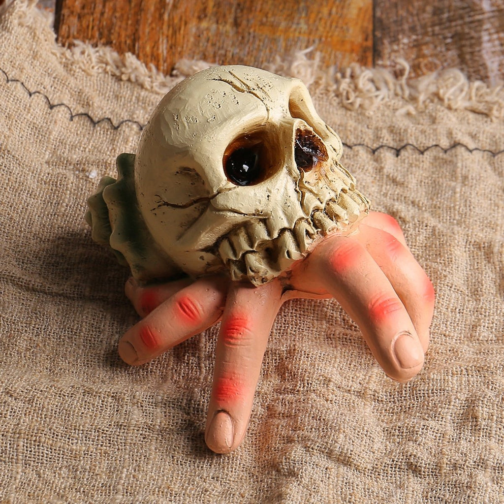 A Halloween prop of a human hand gripping a realistic skull, resting on a textured beige burlap fabric background.