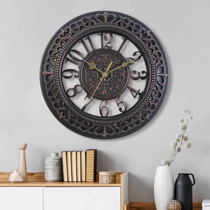Decorative wall clock with intricate design on a gray wall, with books and vases on a shelf below.