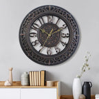 Decorative wall clock with intricate design on a gray wall, with books and vases on a shelf below.