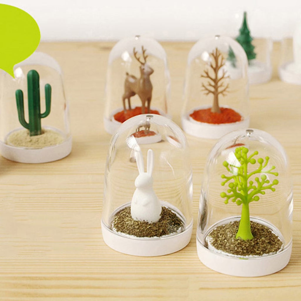 Five glass dome miniatures with rabbit, cactus, deer, and tree figurines on a wooden table, with a green speech bubble and small decor in the background.