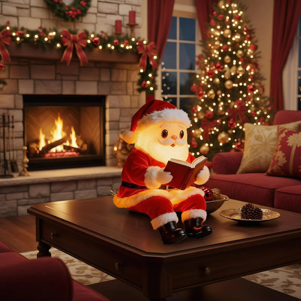 A Santa Claus figure sits on a dark wooden table in a cozy, decorated living room, reading a book. The background features a lit fireplace, a Christmas tree, and a red sofa.