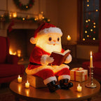 A glowing Santa Claus figure in a red suit sits on a table holding an open book in a warm, candle-lit holiday room. A wrapped gift box, a candlestick with a lit candle, and other candles are arranged around him, with a fireplace and window in the background.
