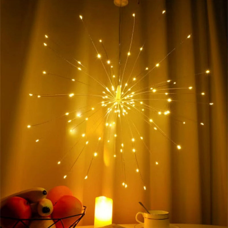 A decorative fiber optic starburst string light with many thin glowing wires radiating outward, shown hanging in a warm yellow-lit room with curtains, a small candle, and a cup on a table.