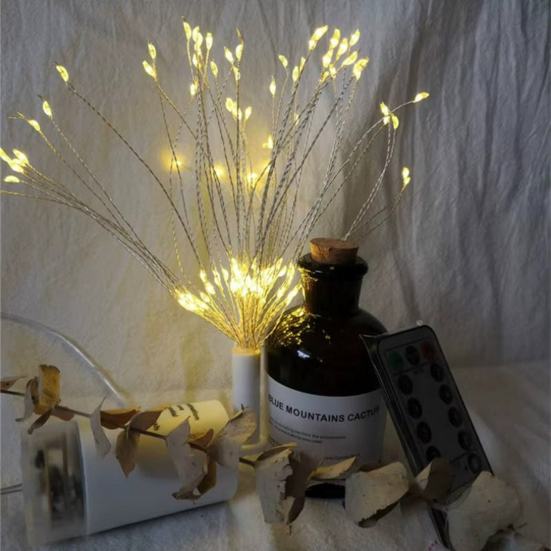 A decorative fiber-optic LED string light set with many thin glowing wires, styled beside an amber bottle, a white diffuser, dried leaves, and a remote control against a textured white fabric background.