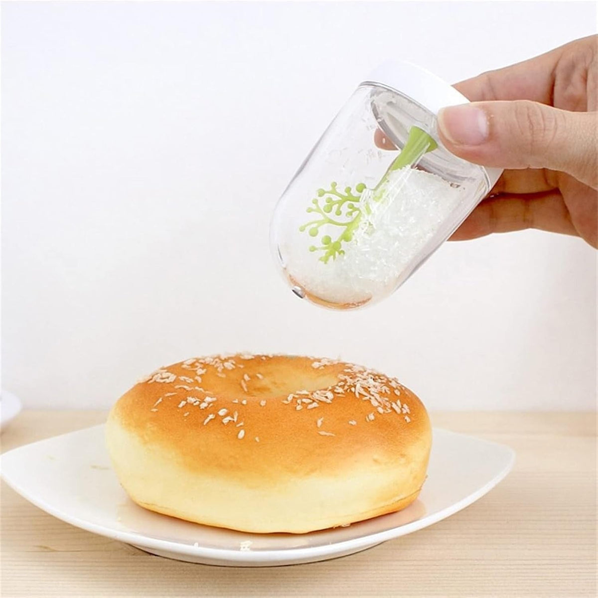A hand holds a clear plastic sprinkler above a sesame-topped bun on a white plate, with white sprinkles falling onto the bun against a clean white background.
