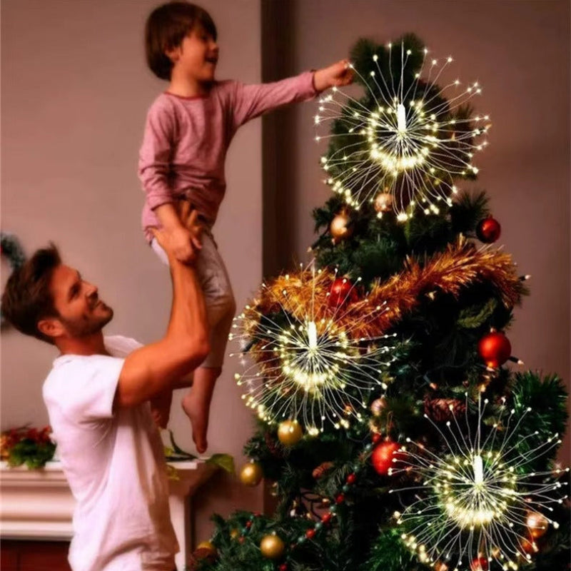 A man and a child decorate a Christmas tree with glowing fiber-optic firework string lights. The warm white lights create bright starburst patterns against the dark green tree, with gold and red ornaments and metallic gold garland adding festive accents.