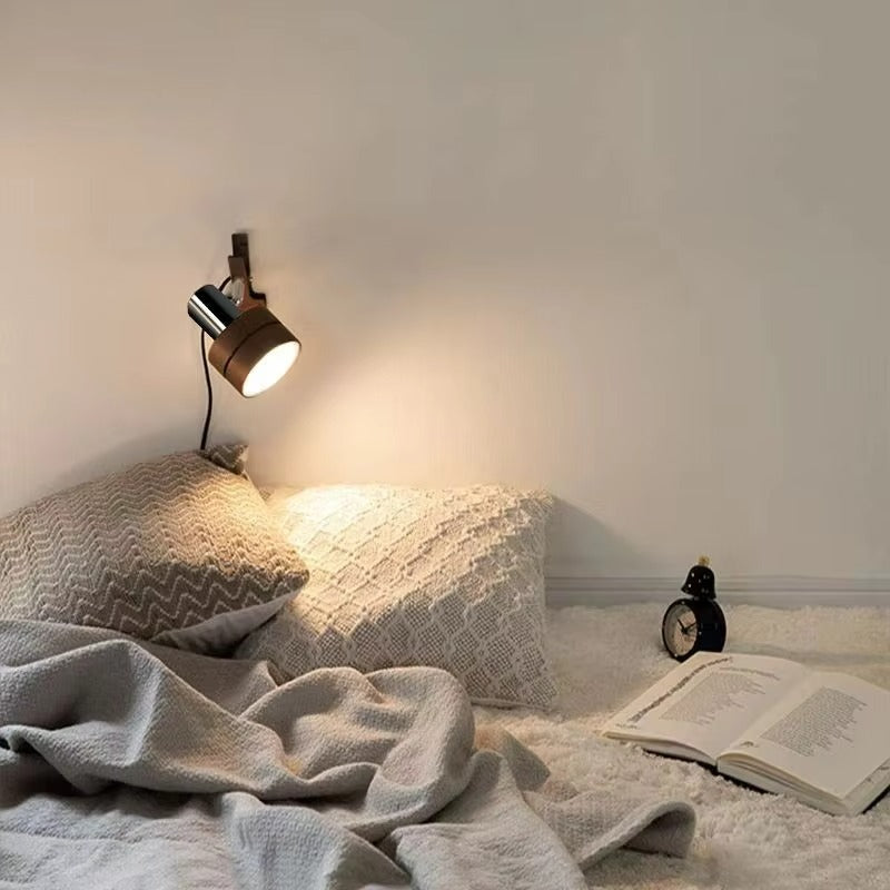 A wall-mounted spotlight with a warm wood-look shade and black metal base, casting light onto a bed with textured pillows, a blanket, and an open book in a softly lit bedroom.