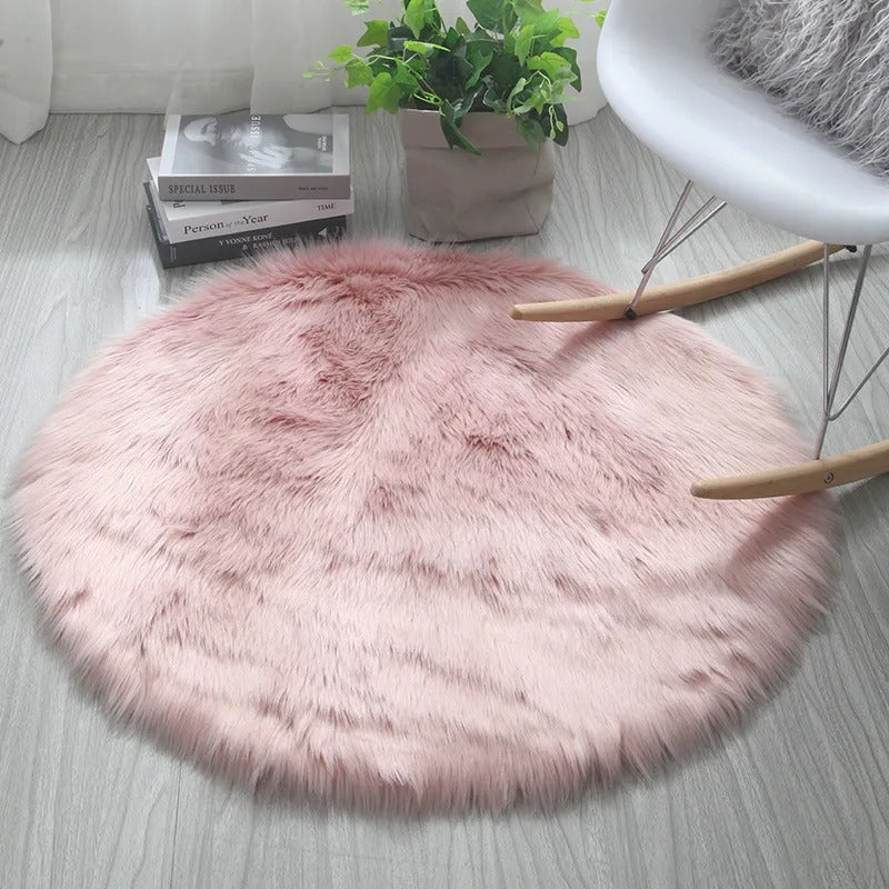 Round pink fluffy rug on a light wooden floor with a plant and books in the background.