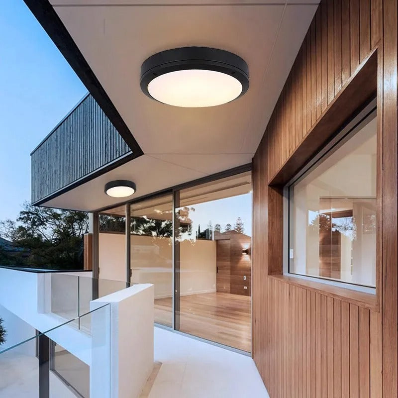 Modern circular flush-mount ceiling light fixture with a black rim and warm white glowing light, installed on a contemporary house exterior with wood paneling and glass sliding doors.