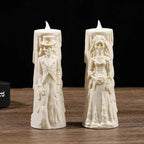 Two cream-colored skeleton wedding candles standing upright on a wooden surface against a black background. One candle depicts a skeleton groom in a suit and top hat, and the other depicts a skeleton bride in a wedding gown holding a bouquet.