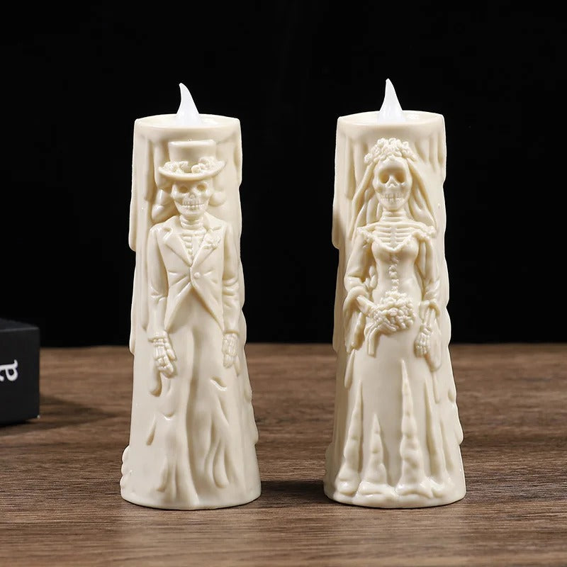 Two cream-colored skeleton wedding candles standing upright on a wooden surface against a black background. One candle depicts a skeleton groom in a suit and top hat, and the other depicts a skeleton bride in a wedding gown holding a bouquet.