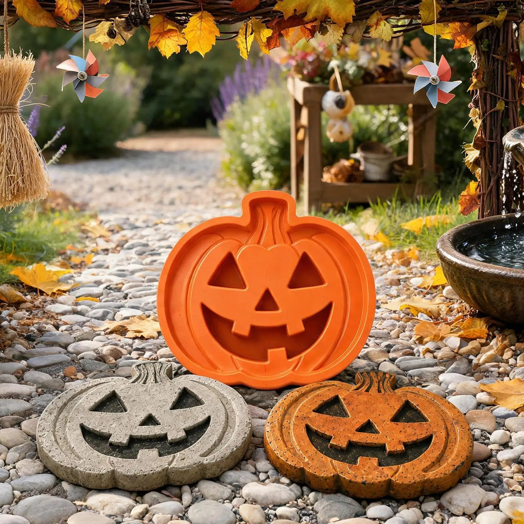 Three pumpkin-shaped decorative items displayed outdoors on a pebble garden path: a bright orange raised jack-o'-lantern in the center, a gray stone jack-o'-lantern in the lower left, and a brown stone jack-o'-lantern in the lower right. Fall leaves, hanging pinwheel ornaments, and a garden setting are visible in the background.