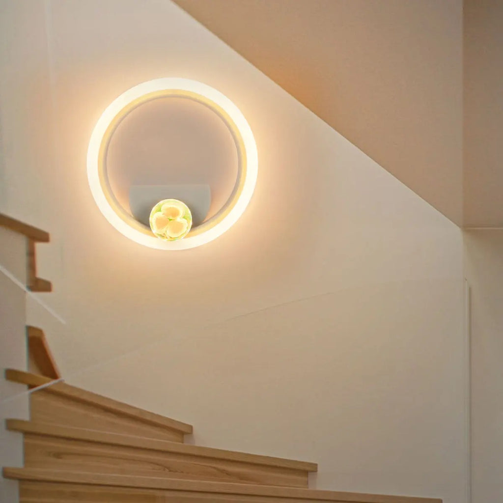 A modern circular wall sconce light fixture with a glowing warm white LED ring and a small central gold-toned decorative element, mounted on a beige wall above wooden stairs.