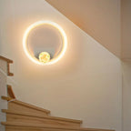 A modern circular wall sconce light fixture with a glowing warm white LED ring and a small central gold-toned decorative element, mounted on a beige wall above wooden stairs.