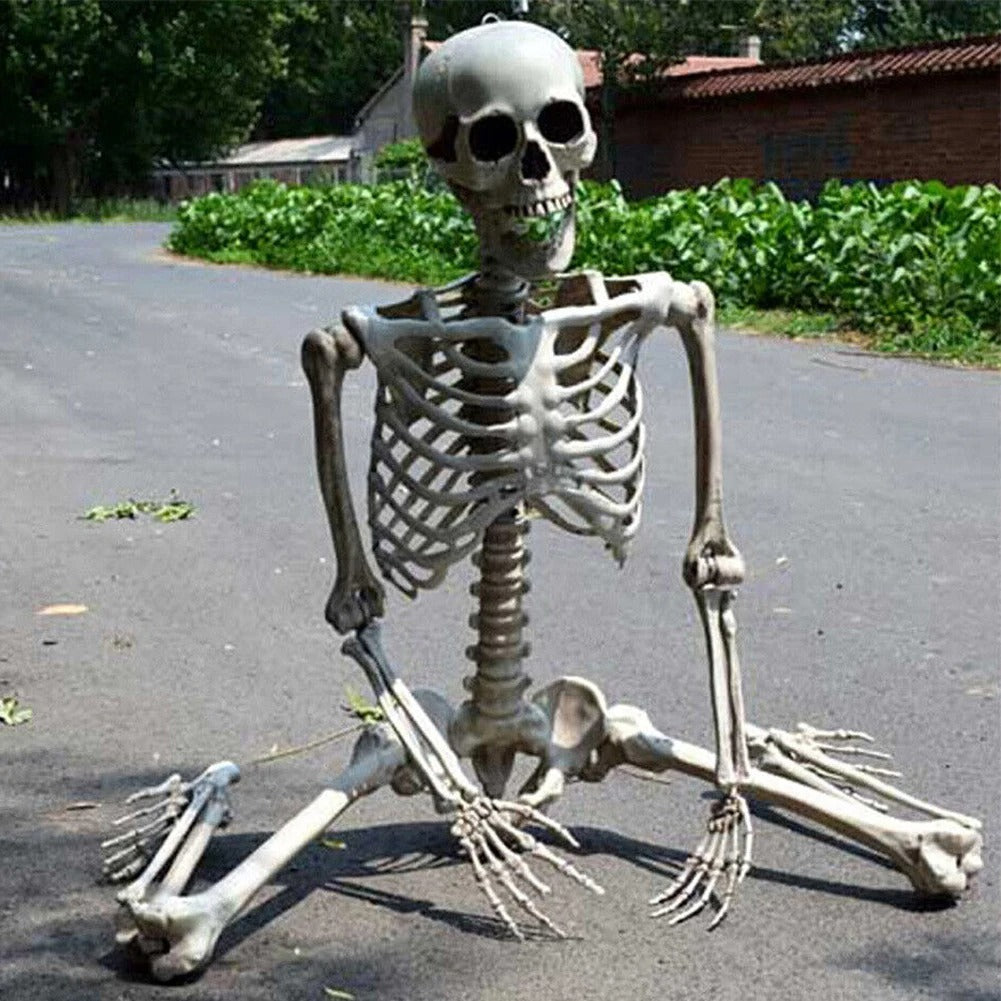 A life-size kneeling plastic skeleton decoration sitting on an asphalt road outdoors with greenery and buildings in the background.
