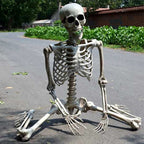 A life-size kneeling plastic skeleton decoration sitting on an asphalt road outdoors with greenery and buildings in the background.
