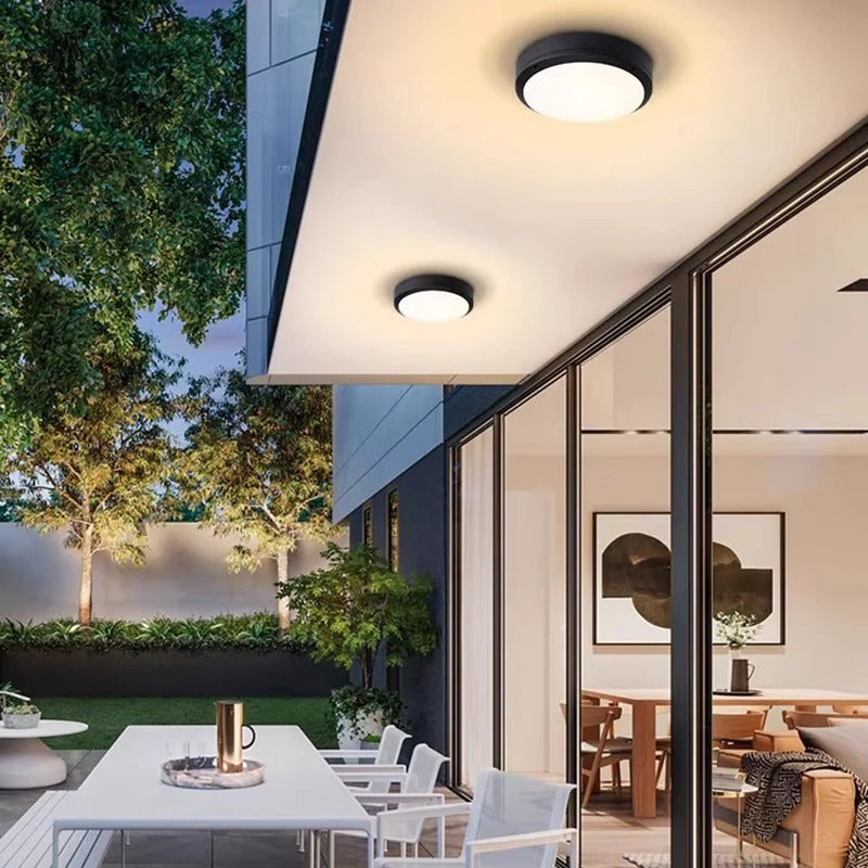 Modern black circular flush-mount ceiling lights installed on a white overhang of a contemporary glass-walled building, with an outdoor patio and dining area visible below at dusk.