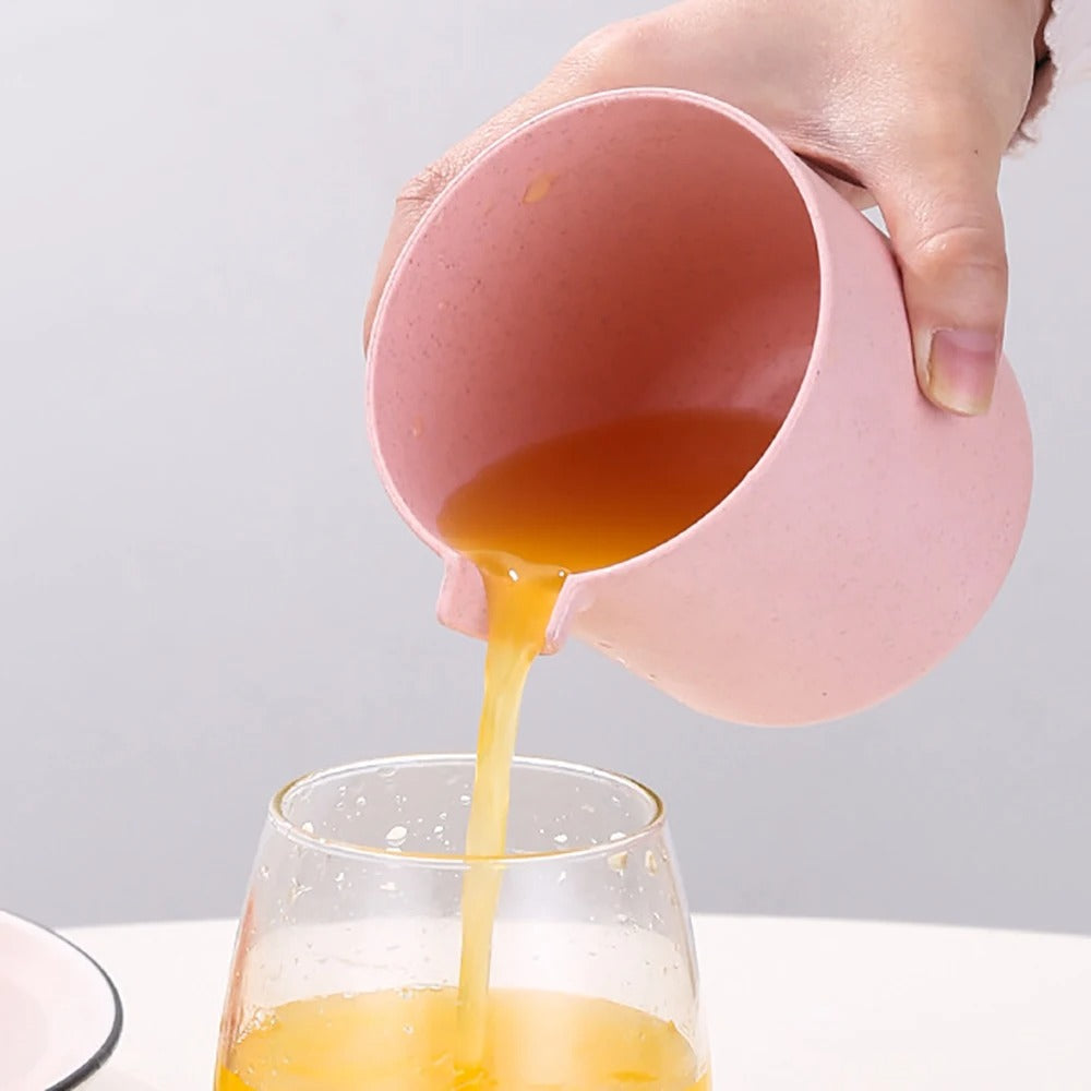 A hand holds a pink pitcher pouring orange juice into a clear glass, shown against a light gray background.