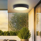 A modern flush-mount circular ceiling light with a black outer frame and a glowing warm white light, installed on a ceiling in a contemporary outdoor patio setting with plants and vertical slats.