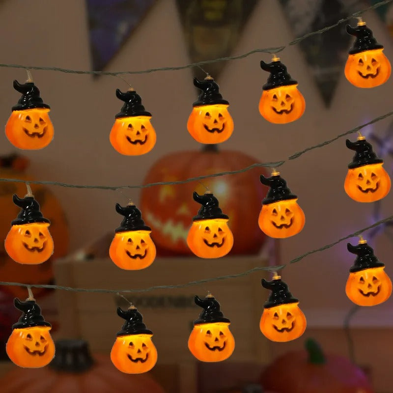 Halloween string lights featuring glowing orange jack-o'-lantern pumpkins wearing black witch hats, shown hanging against a softly blurred indoor background with more pumpkins visible.