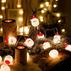 Decorative setup with candles, Santa Claus figurines, and string lights on a wooden surface.