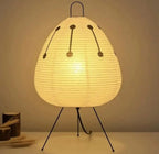 A warm white paper lantern table lamp with a ribbed teardrop shape, supported by three black metal legs, shown lit against a neutral beige background.
