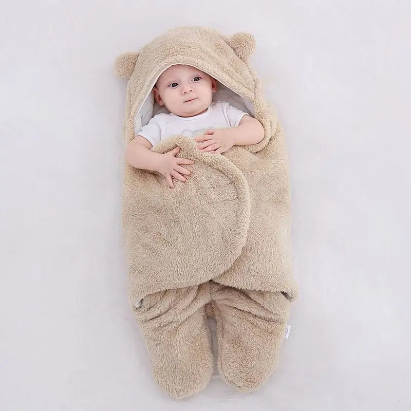 A baby wrapped in a plush beige bear-themed swaddle blanket with a hood featuring bear ears, lying on a light gray background.